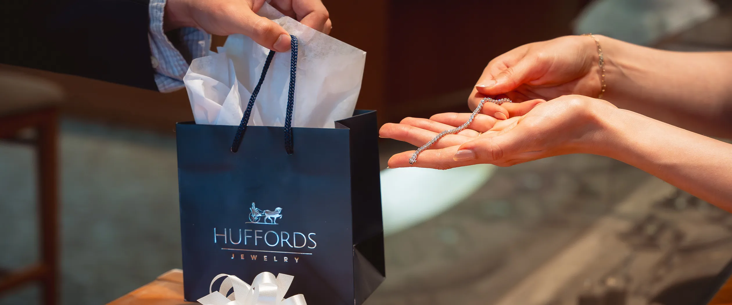 Hands holding a diamond bracelet over a Huffords Jewelry gift bag. Click to contact us.