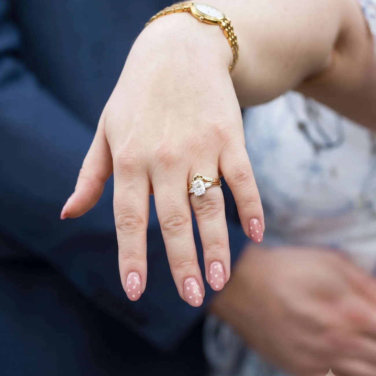 A hand with a gold watch and a diamond ring on the ring finger, nails painted with pink polka dots.