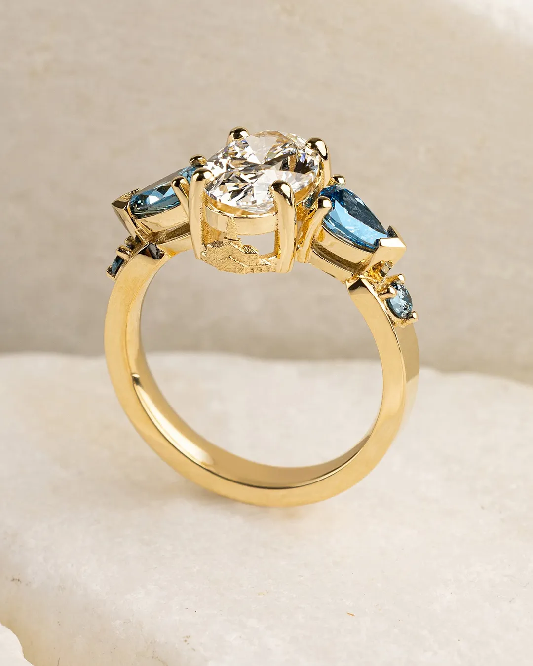 A gold ring with a large central diamond and two blue side stones on a marble surface.