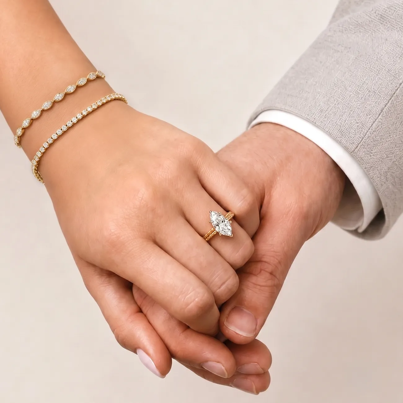 Two hands clasped; one with a marquise diamond ring and gold bracelets, the other in a light gray suit.