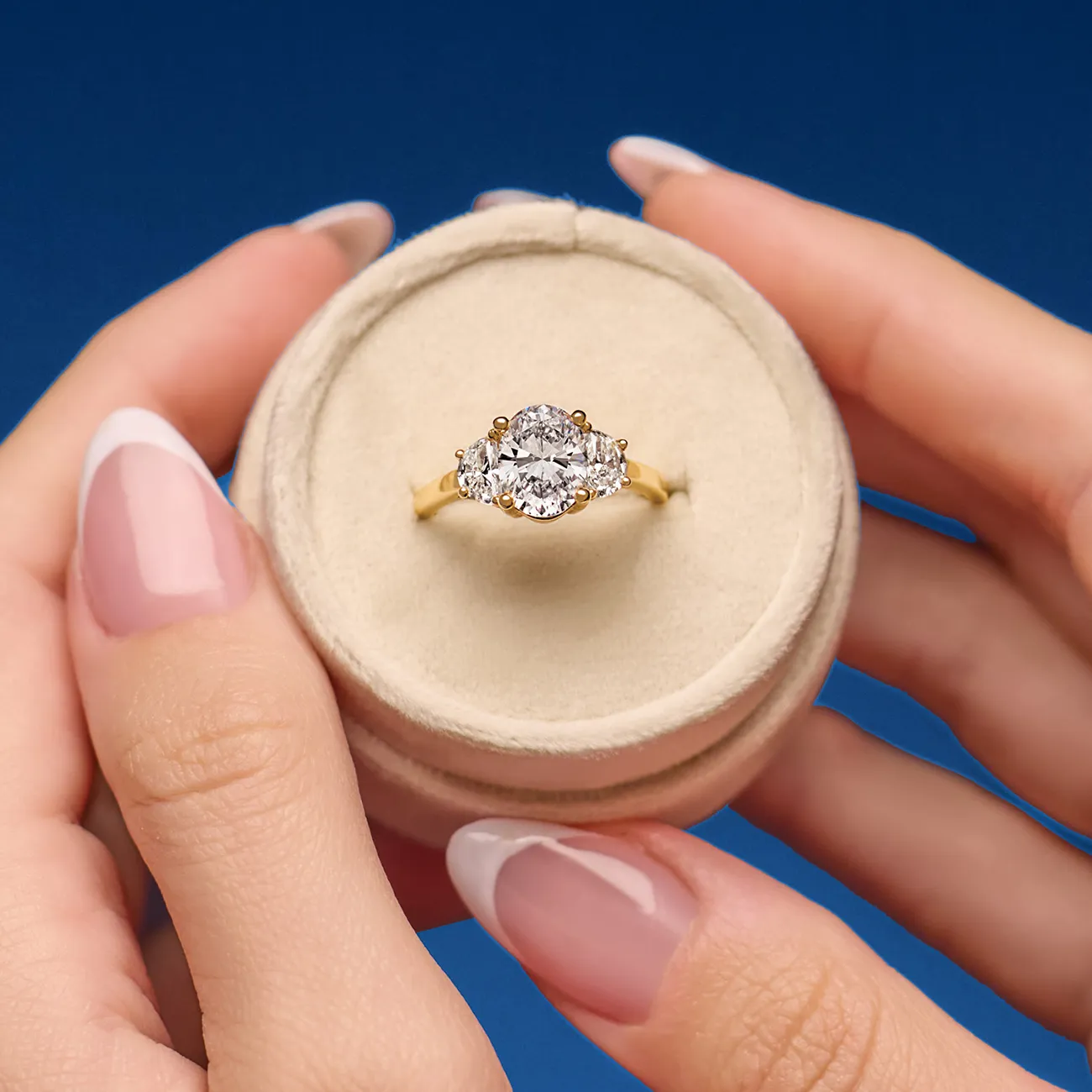 A gold ring with three diamonds in a round box, held by hands with manicured nails.