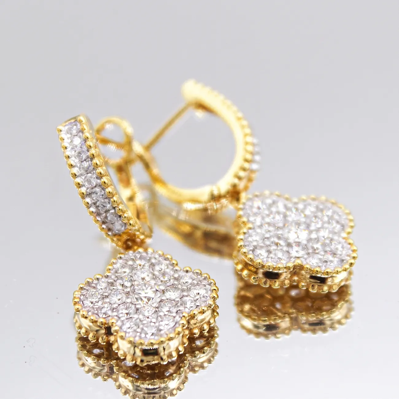 Gold hoop earrings with diamond-studded clover-shaped pendants on a reflective surface.