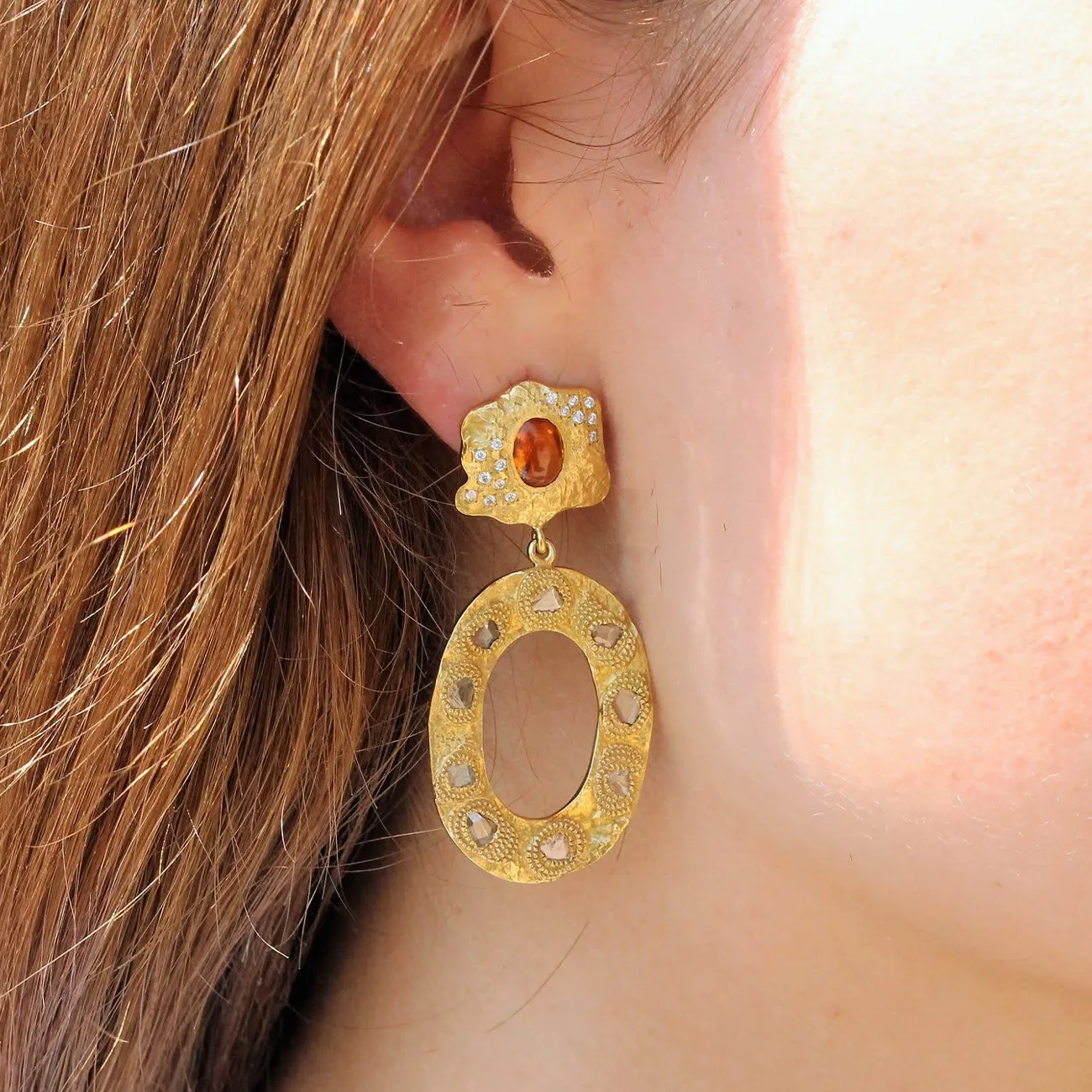 Gold earring with orange gemstone and open oval design worn on ear.