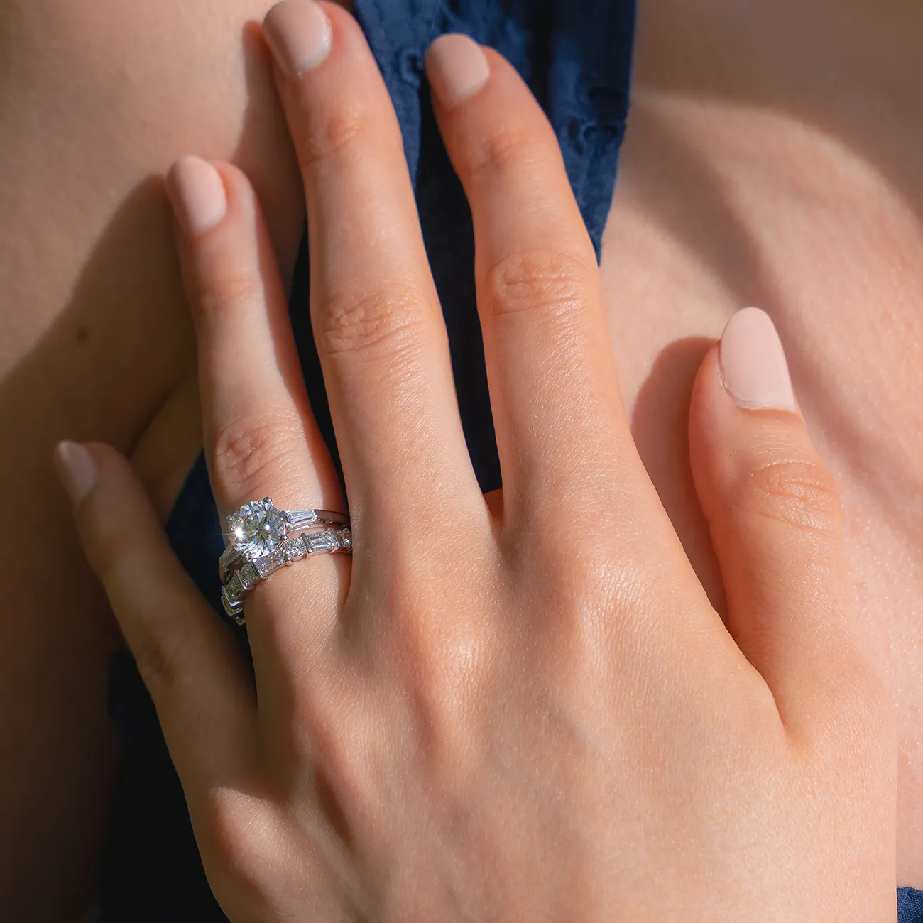 A hand with a diamond engagement ring set against a blue fabric background.