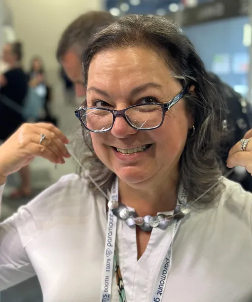 A woman named Angela Peterson holding up a necklace while smiling and wearing glasses.