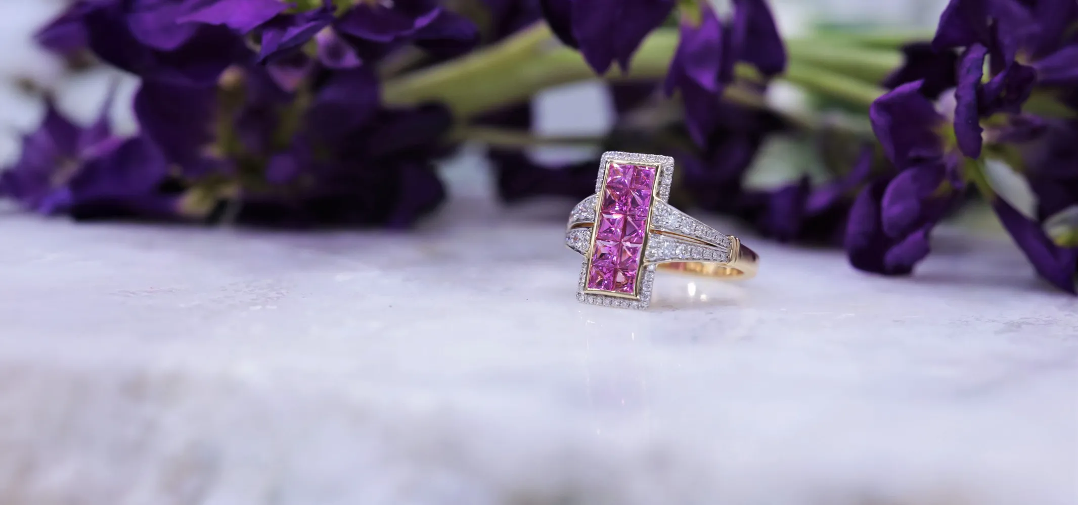 Pink gemstone ring with diamond accents on a marble surface, surrounded by purple flowers. 