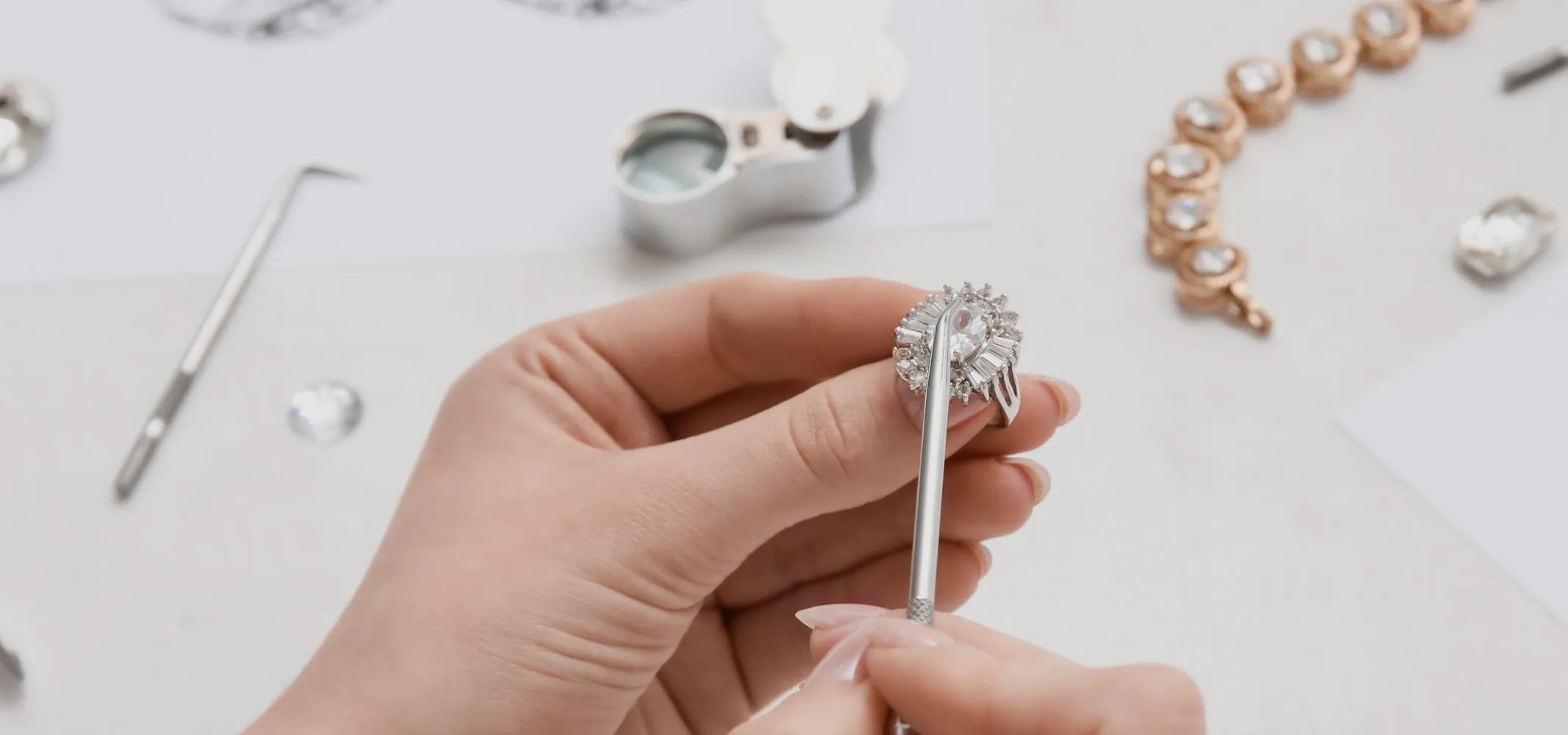 Hand holding a diamond ring with a tool, surrounded by jewelry-making supplies. Click to start a project at Hupp Jewelers.