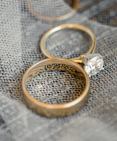 Close-up of two gold rings, one with a diamond, engraved with the date 10/21/2023 on a textured fabric background.
