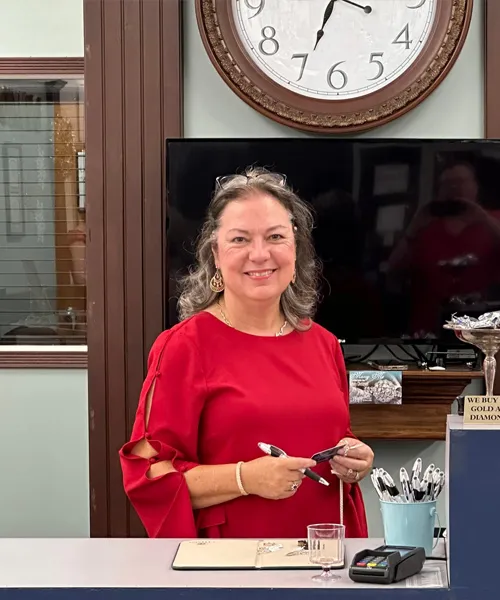 Angela Peterson in a red blouse smiling at a jewelry store counter, holding pens, with a clock and TV in the background.