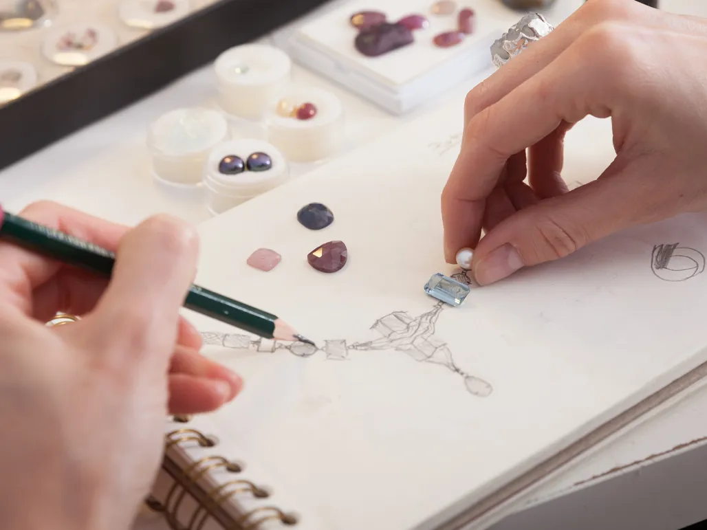 A hand sketches jewelry designs while selecting gemstones, showcasing custom jewelry creation.