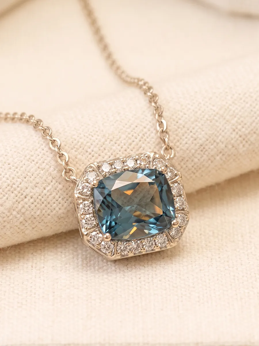 A blue gemstone pendant with diamond accents on a gold chain, displayed on a beige fabric.