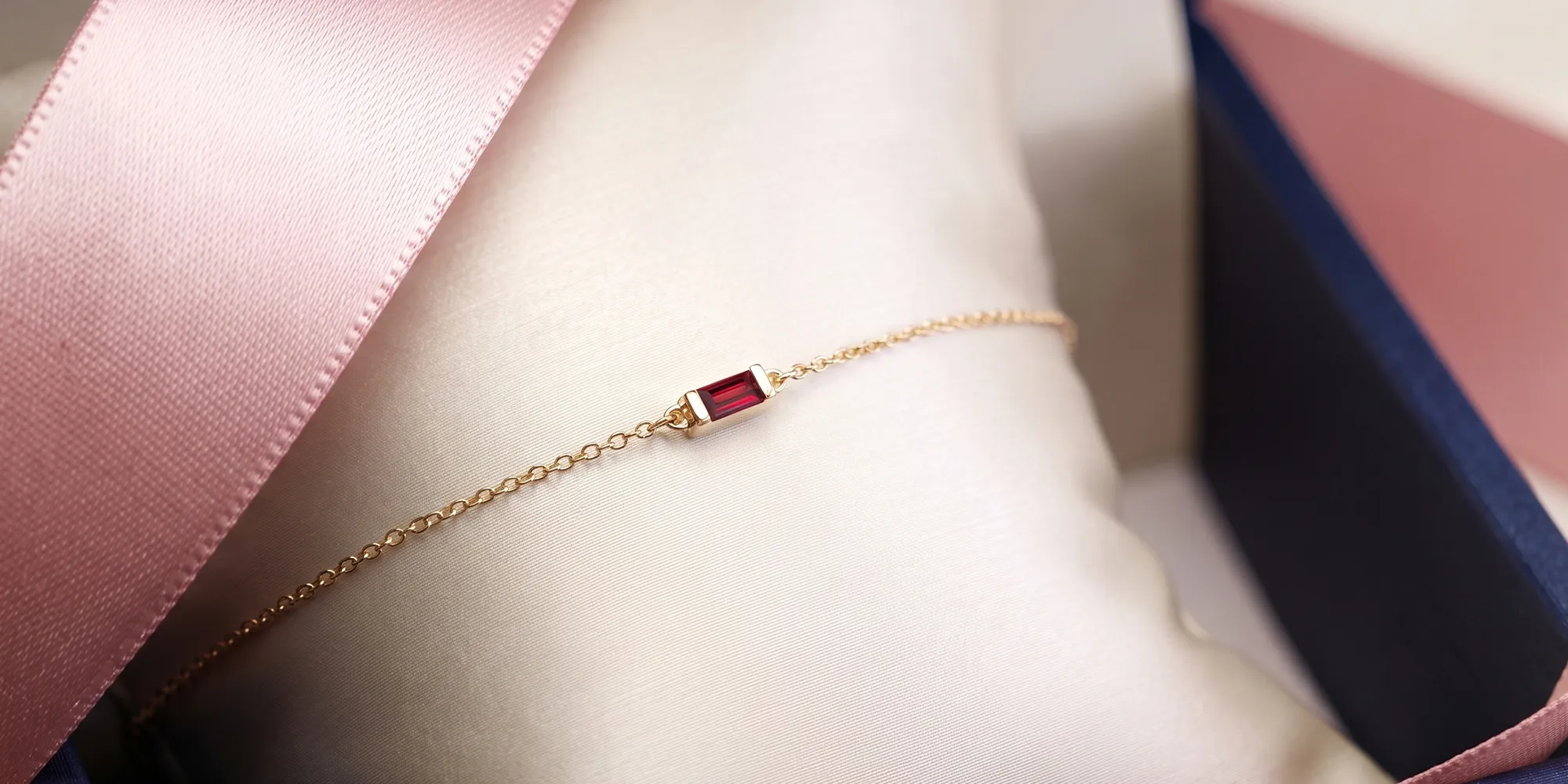 A delicate gold bracelet with a red gemstone on a satin cushion, next to a pink ribbon.