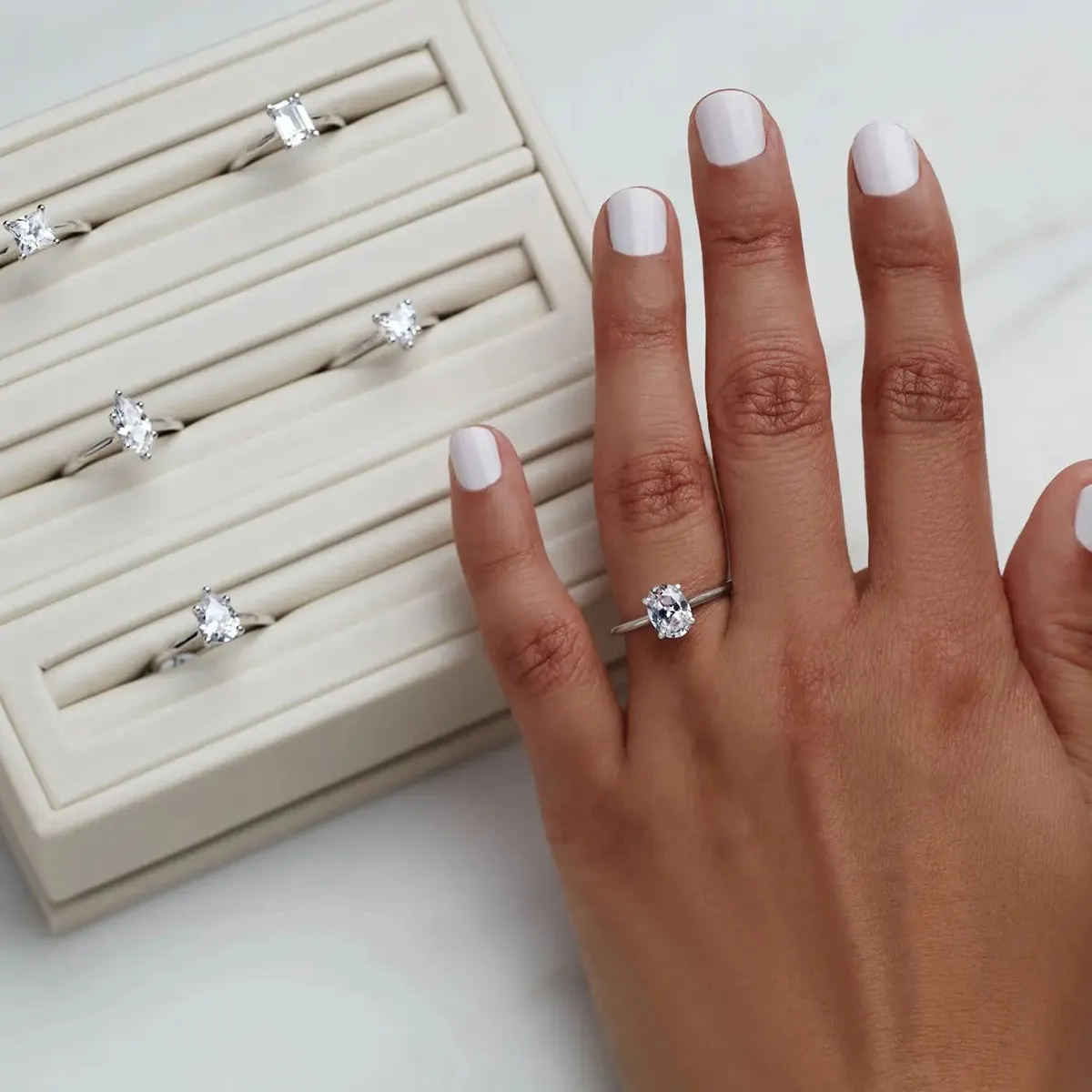 A hand with a white manicure wearing a diamond ring, next to a display of similar rings.