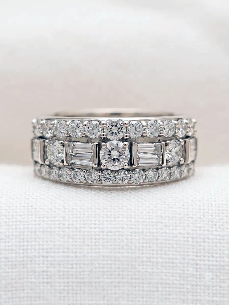 A diamond ring with multiple rows of round and baguette-cut diamonds on a light fabric background.