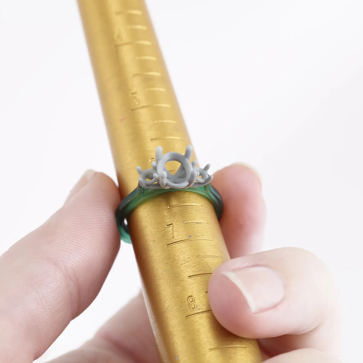 A hand holds a ring with a gray wax setting on a gold ring sizer.