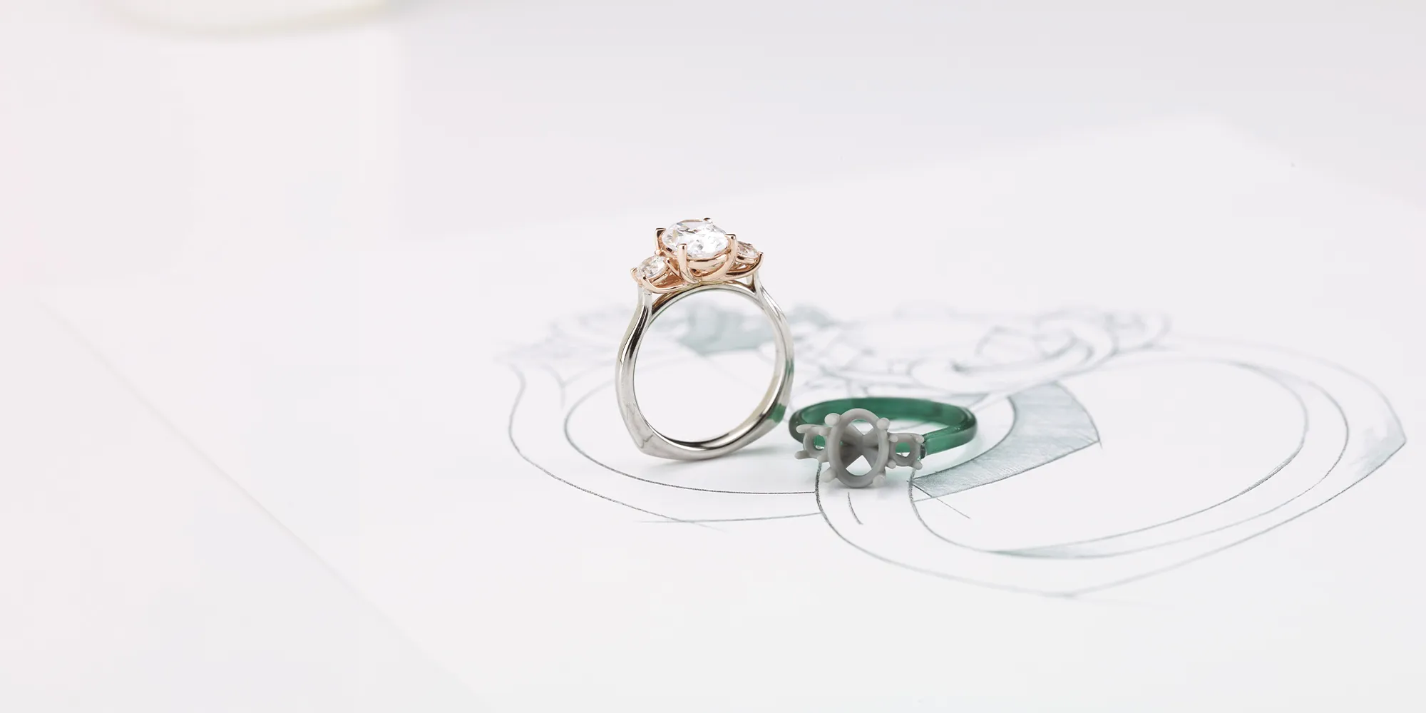 A silver and rose gold engagement ring with a diamond next to a green ring mold on a sketch.