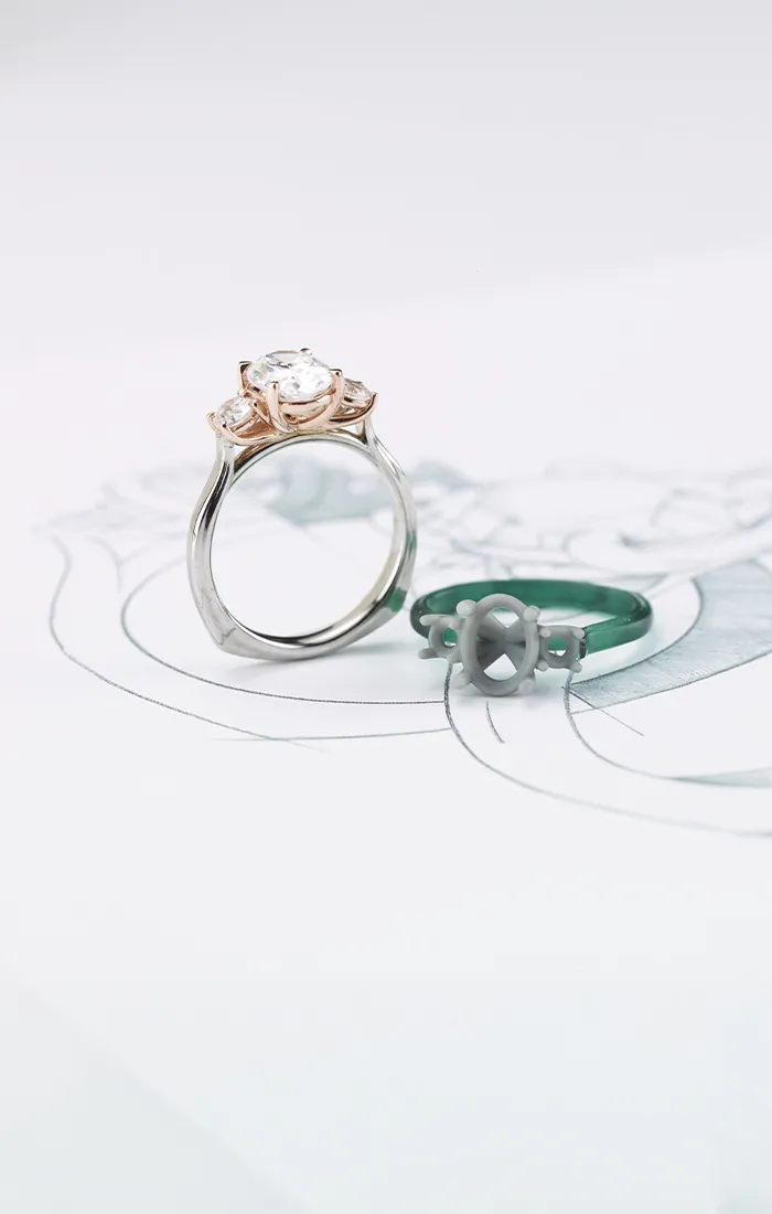 A silver and rose gold engagement ring with a diamond next to a green ring mold on a sketch.