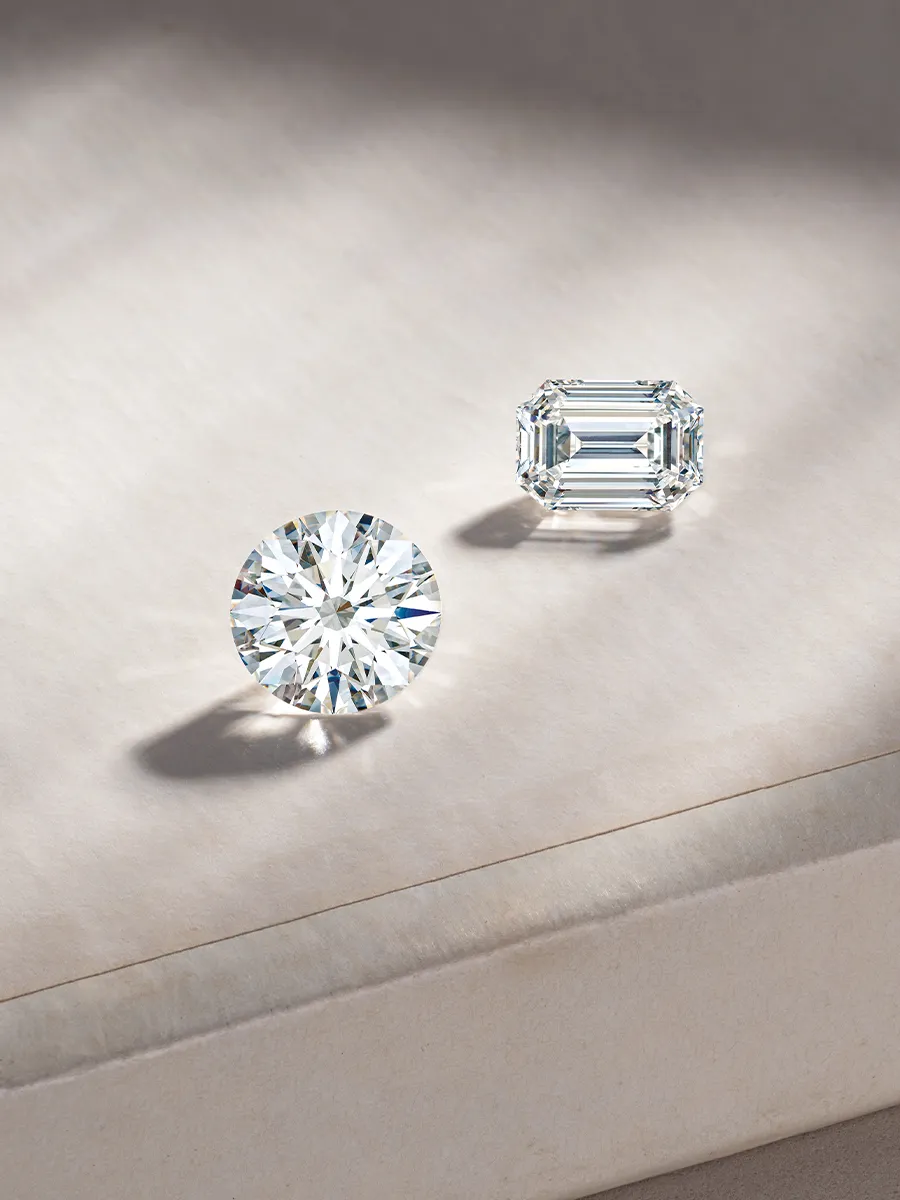 Two diamonds on a soft surface; one round and one emerald cut, both sparkling under light.