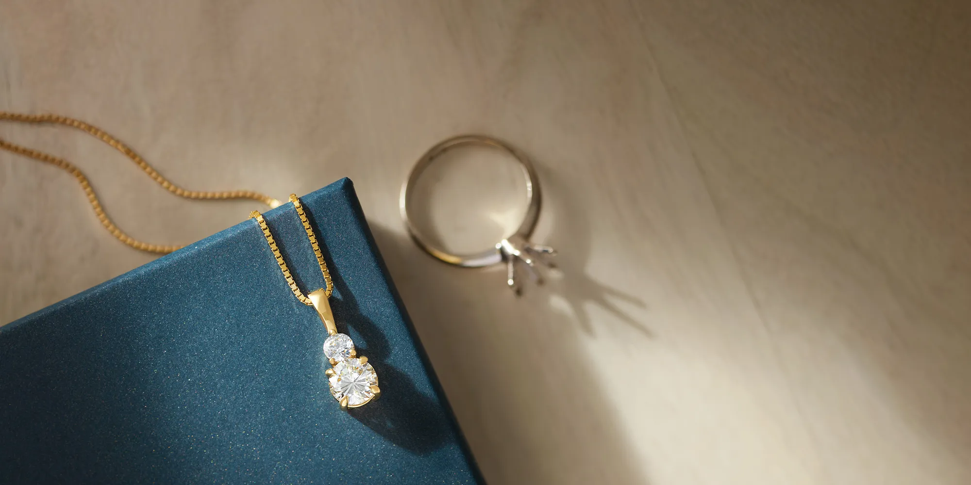 A gold necklace with two diamonds on a blue box, next to a ring on a wooden surface.