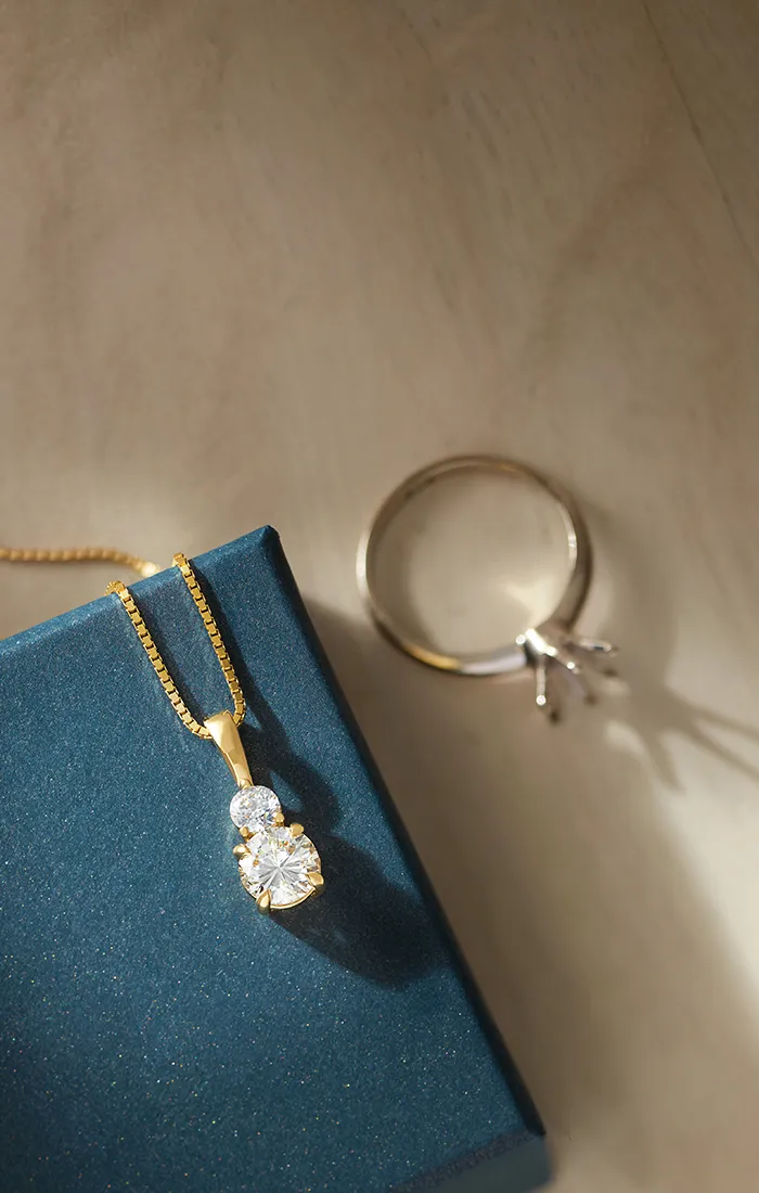 A gold necklace with two diamonds on a blue box, next to a ring on a wooden surface.