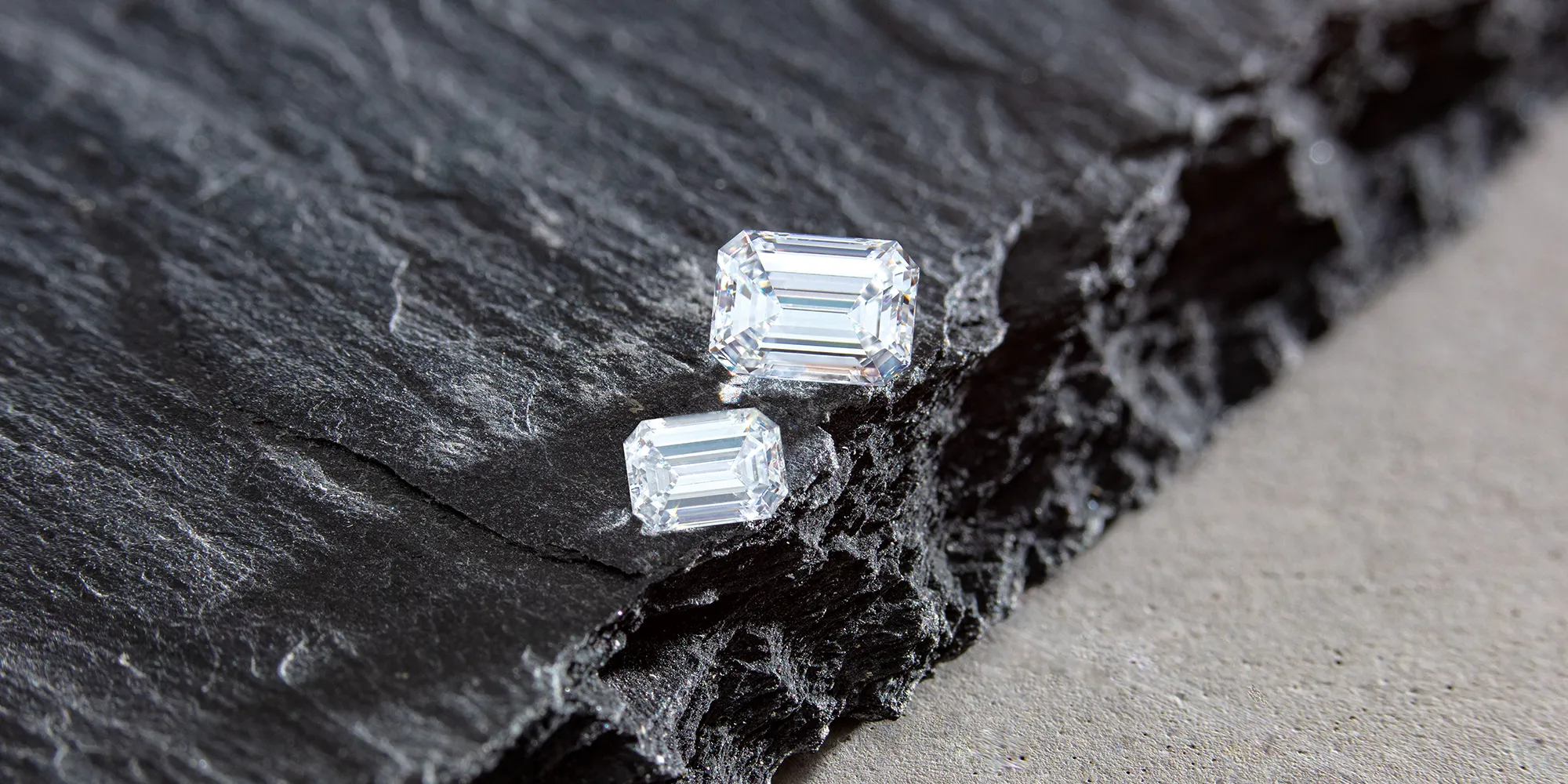 Two emerald-cut diamonds on a textured dark slate surface.