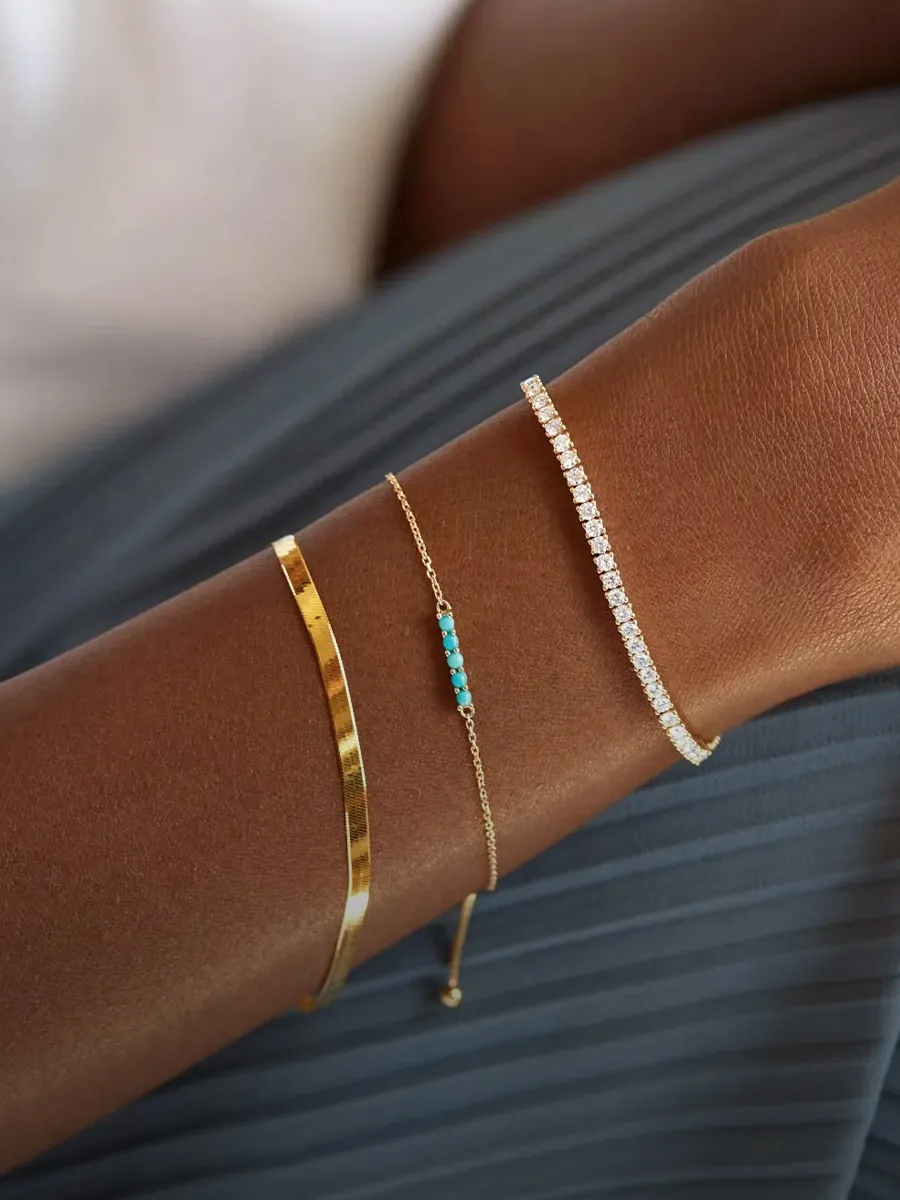Three elegant bracelets on an arm: a gold bangle, a turquoise beaded chain, and a diamond tennis bracelet.