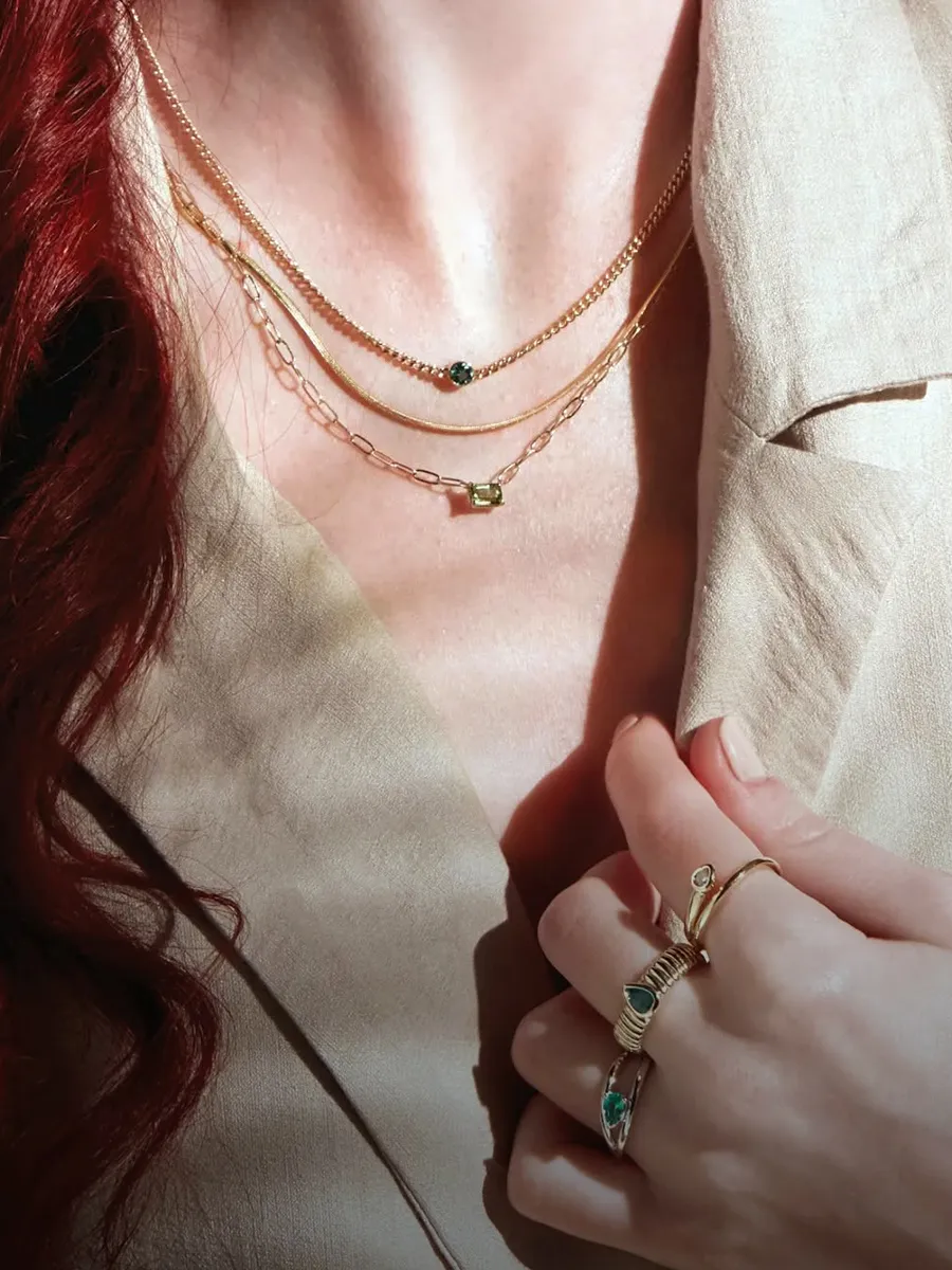 A person wearing layered gold necklaces with green stones and matching rings on a beige outfit.