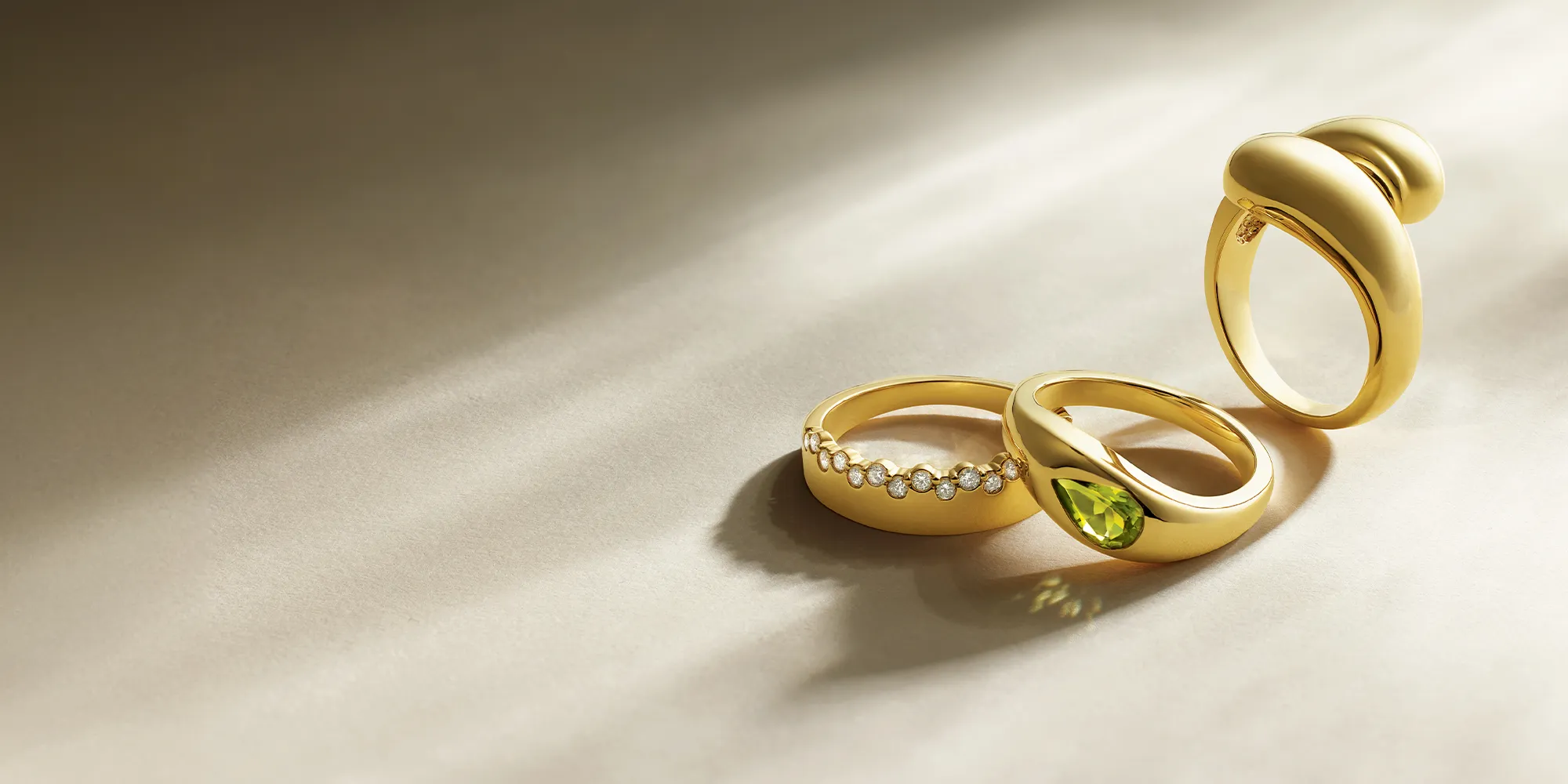 Three gold rings on a light surface; one with diamonds, one with a green gemstone, and one with a bold design.