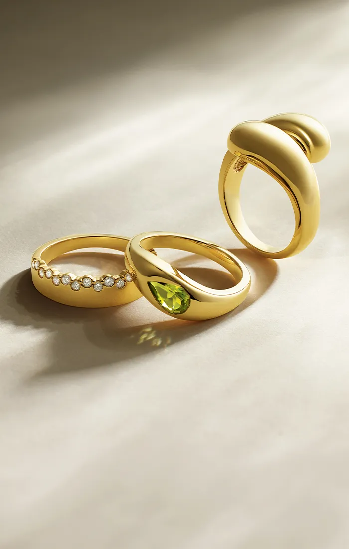 Three gold rings on a light surface; one with diamonds, one with a green gemstone, and one with a bold design.