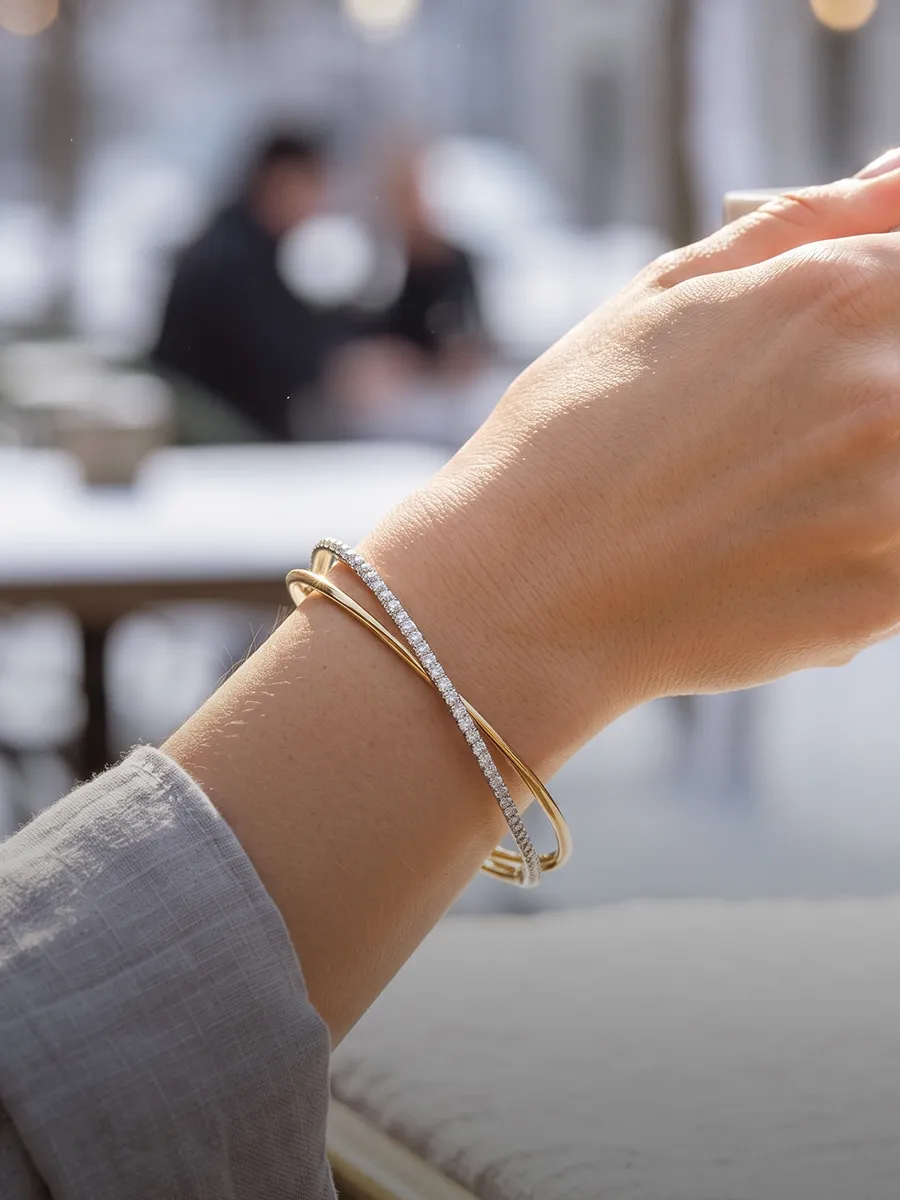 A hand wearing two elegant bracelets, one gold and one with diamonds, in an outdoor setting.