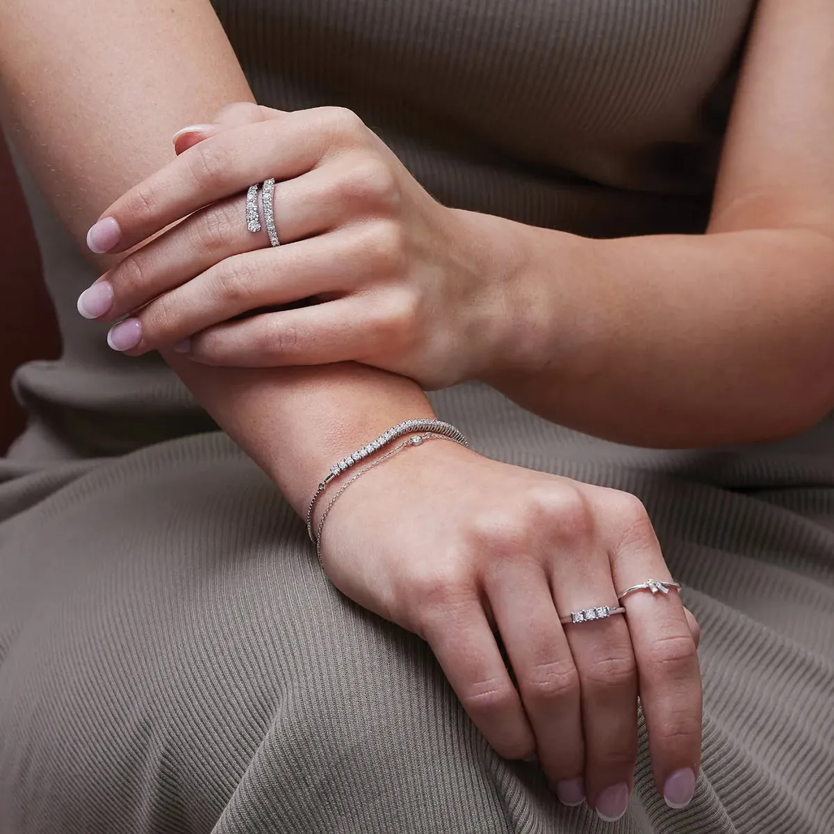 A person wearing diamond rings and a bracelet on their hands, resting on a gray dress.