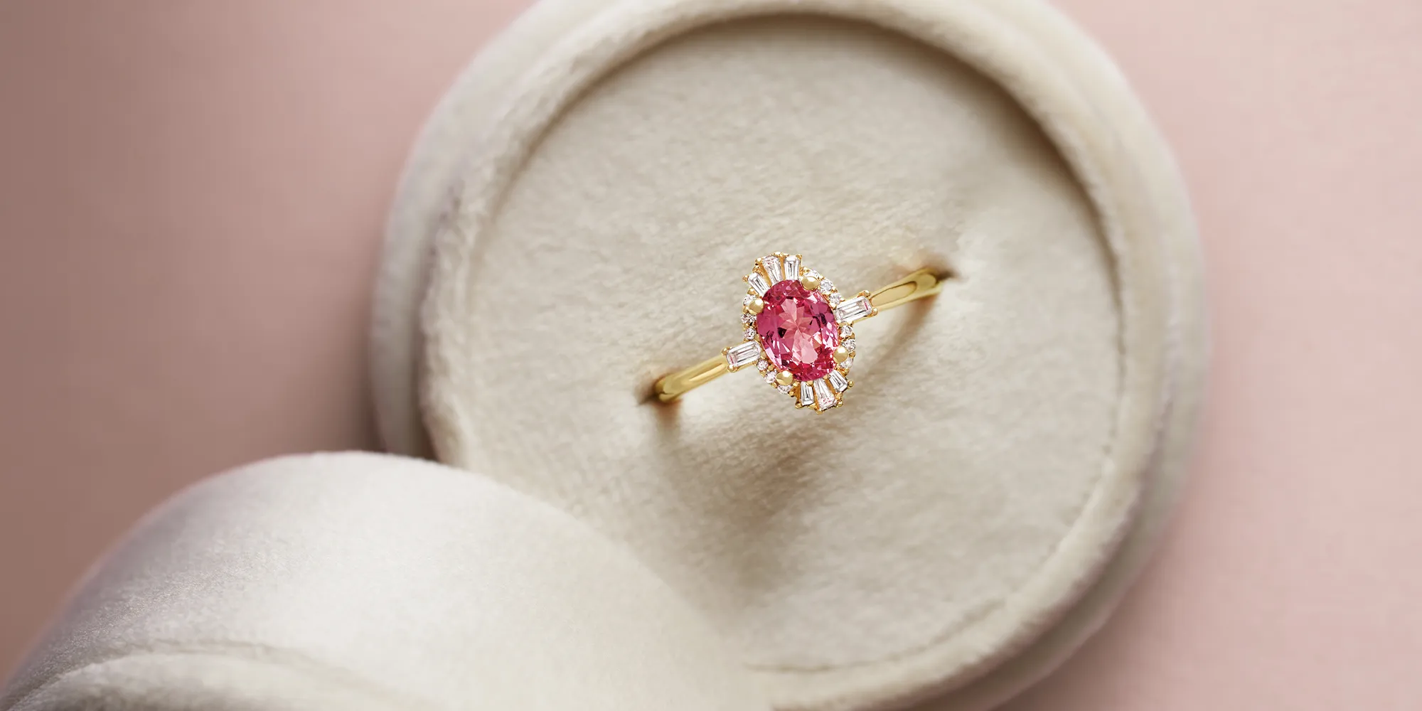 A gold ring with a pink gemstone and diamond accents in a round beige jewelry box.