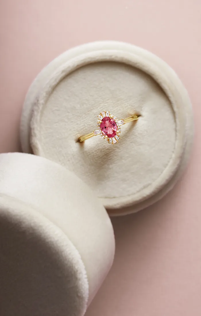 A gold ring with a pink gemstone and diamond accents in a round beige jewelry box.