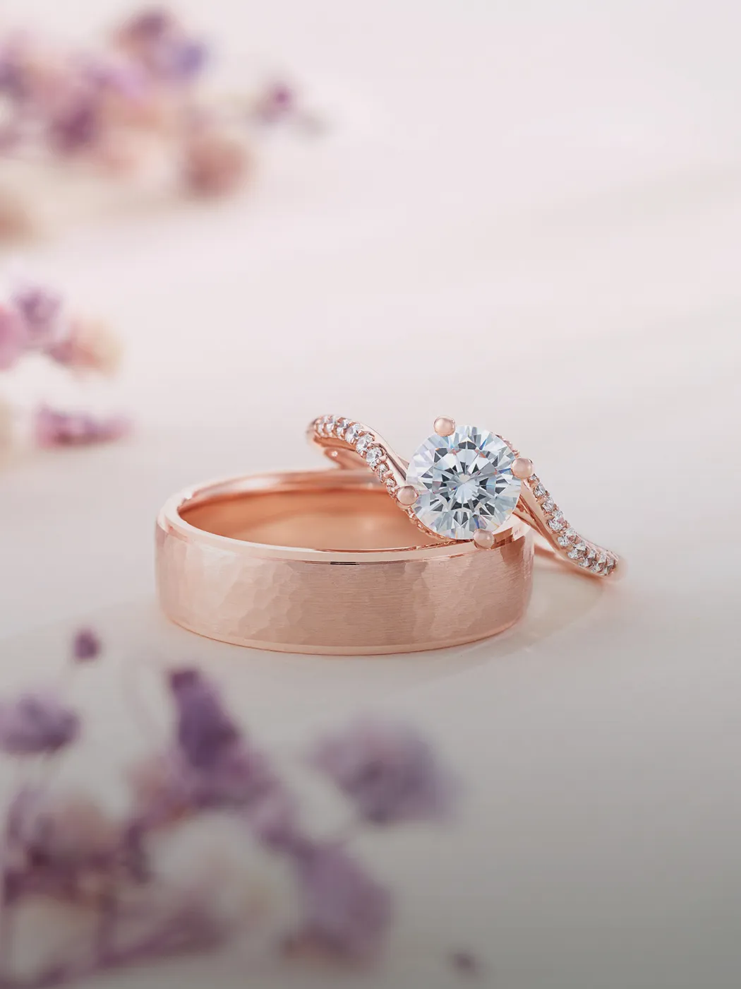 Rose gold engagement and wedding rings with a diamond, surrounded by soft-focus flowers.