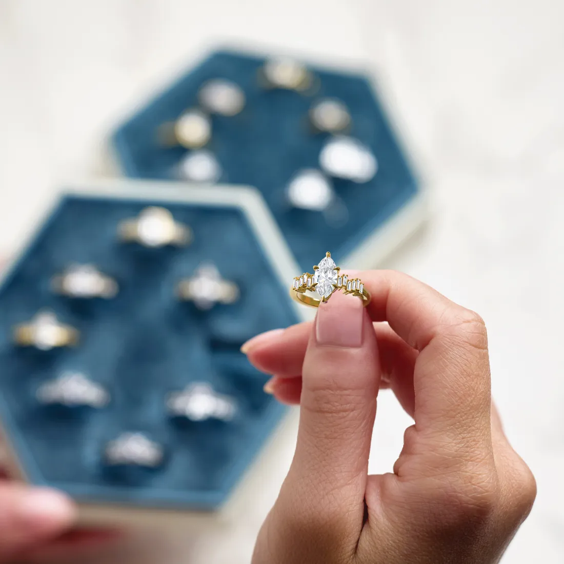 A hand holds a gold engagement ring with a marquise diamond, with more rings in a blue velvet display in the background.
