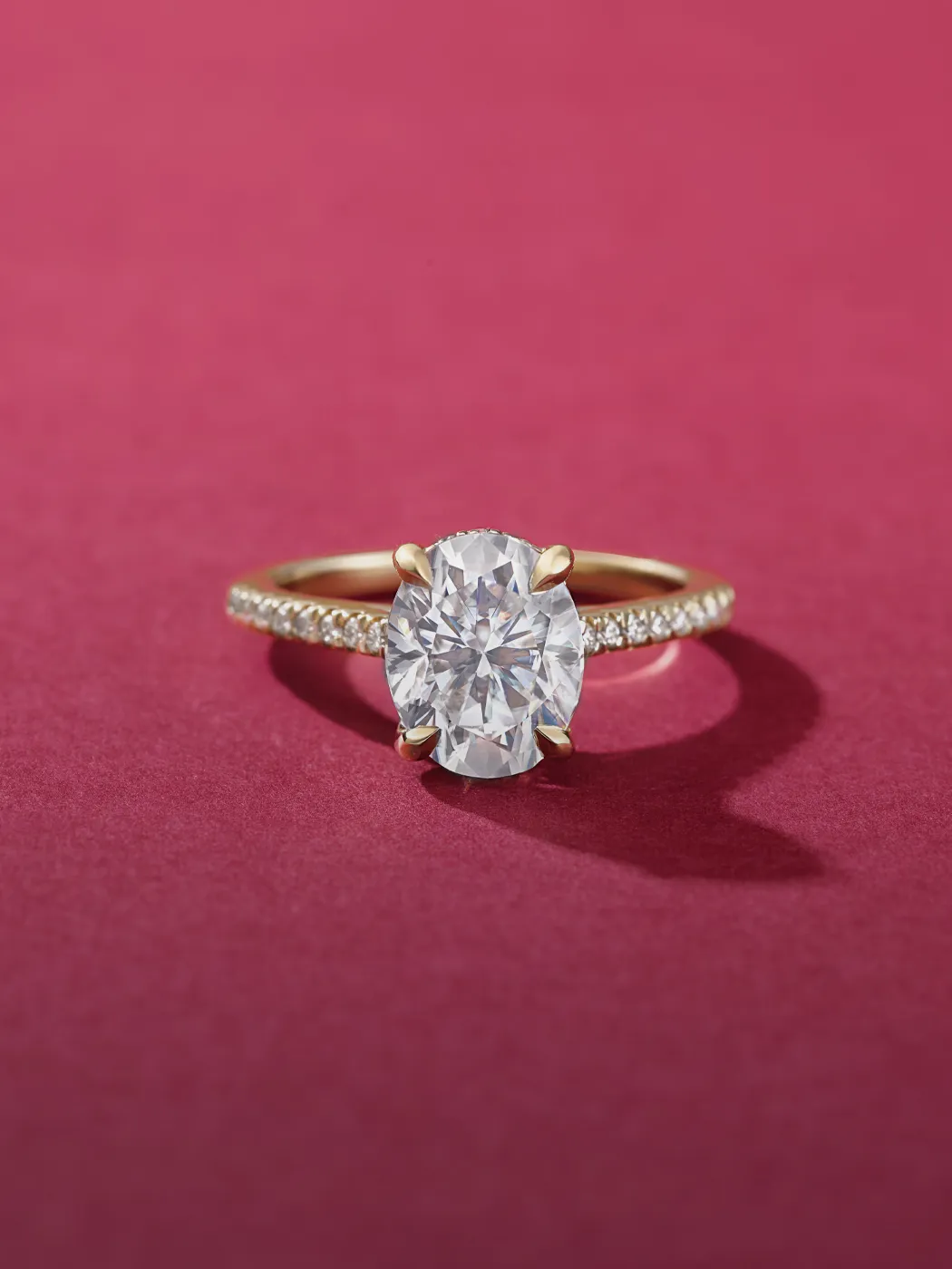 Gold ring with large oval diamond and pave band on a pink background.