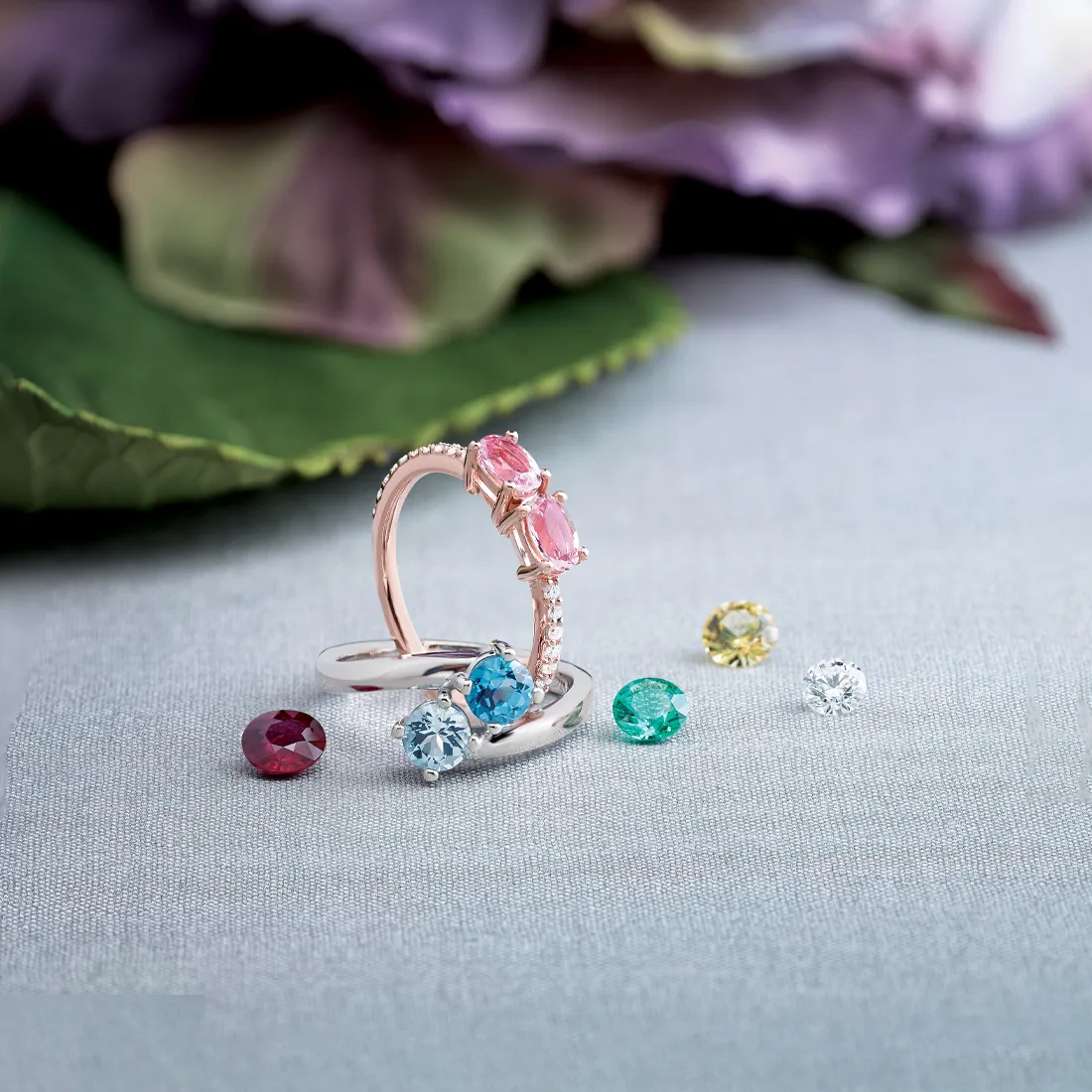 Rings with pink, blue, and clear gemstones on a fabric surface with scattered gems.