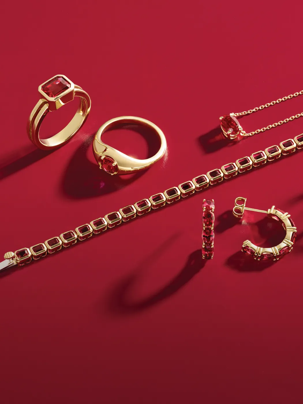 Gold jewelry with red gemstones on a red background, including rings, a bracelet, earrings, and a necklace.