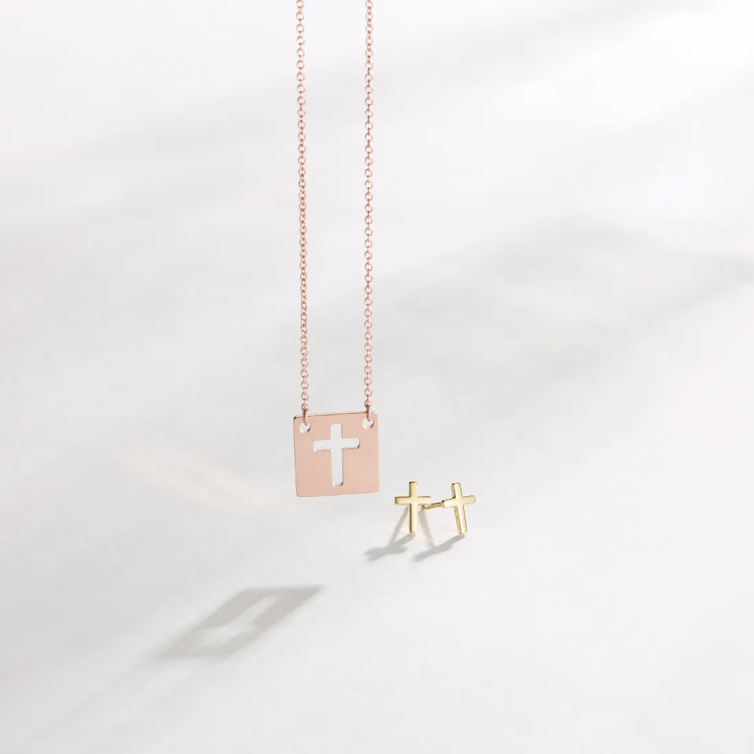 Square pendant necklace with cross cutout and gold cross earrings on a light background.