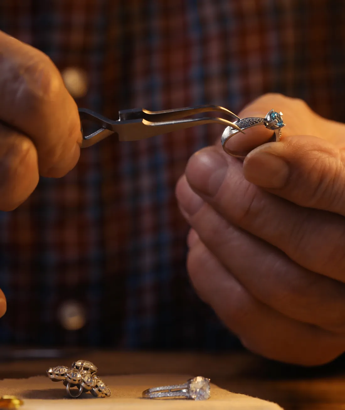 A jeweler uses pliers to handle a ring with a blue gemstone, surrounded by other jewelry pieces.