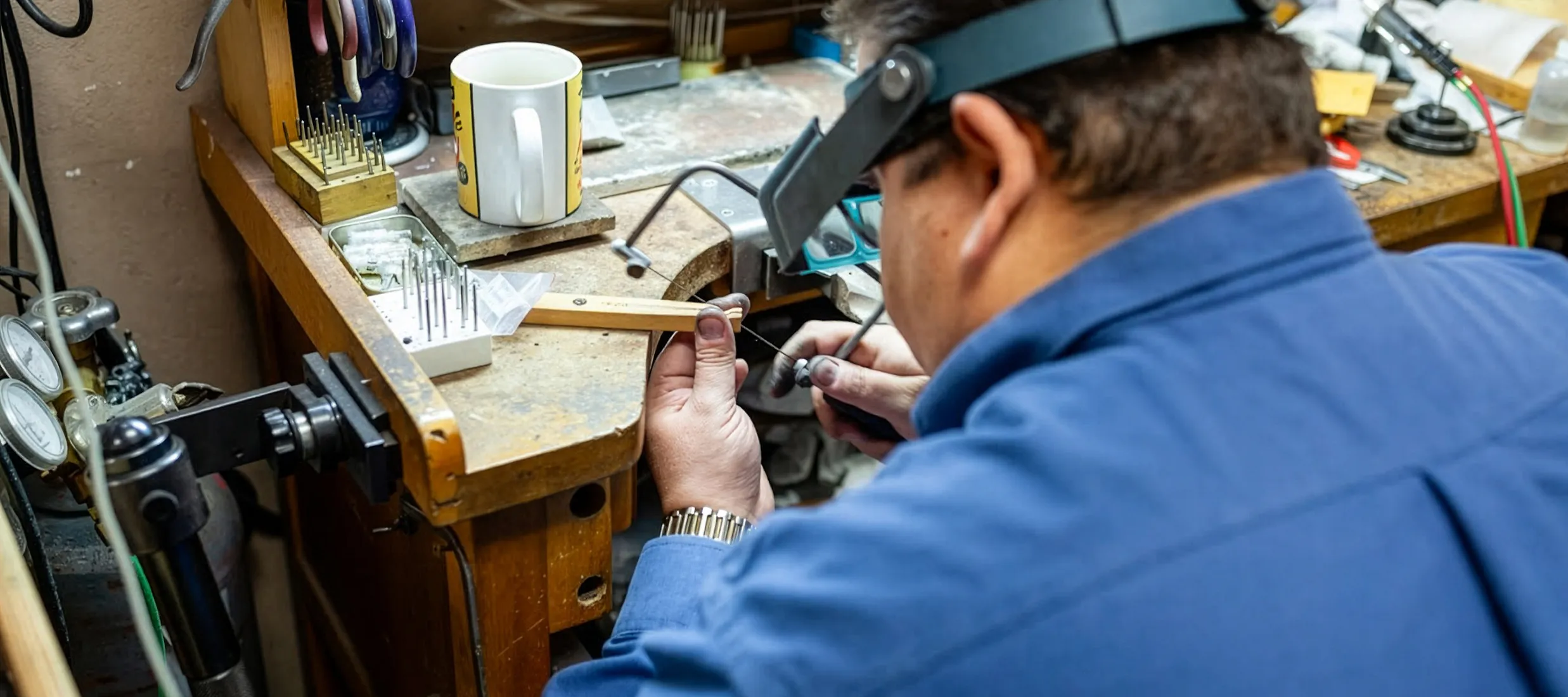 Jeweler working on a piece at a workbench with tools and a coffee mug. Click to view our services at J.A. Jewelers.