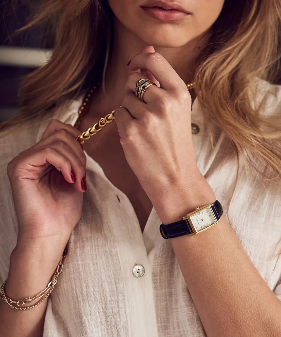 Close-up of a woman's hand wearing a gold chain bracelet, rings, and a vintage watch, styled with a white shirt.