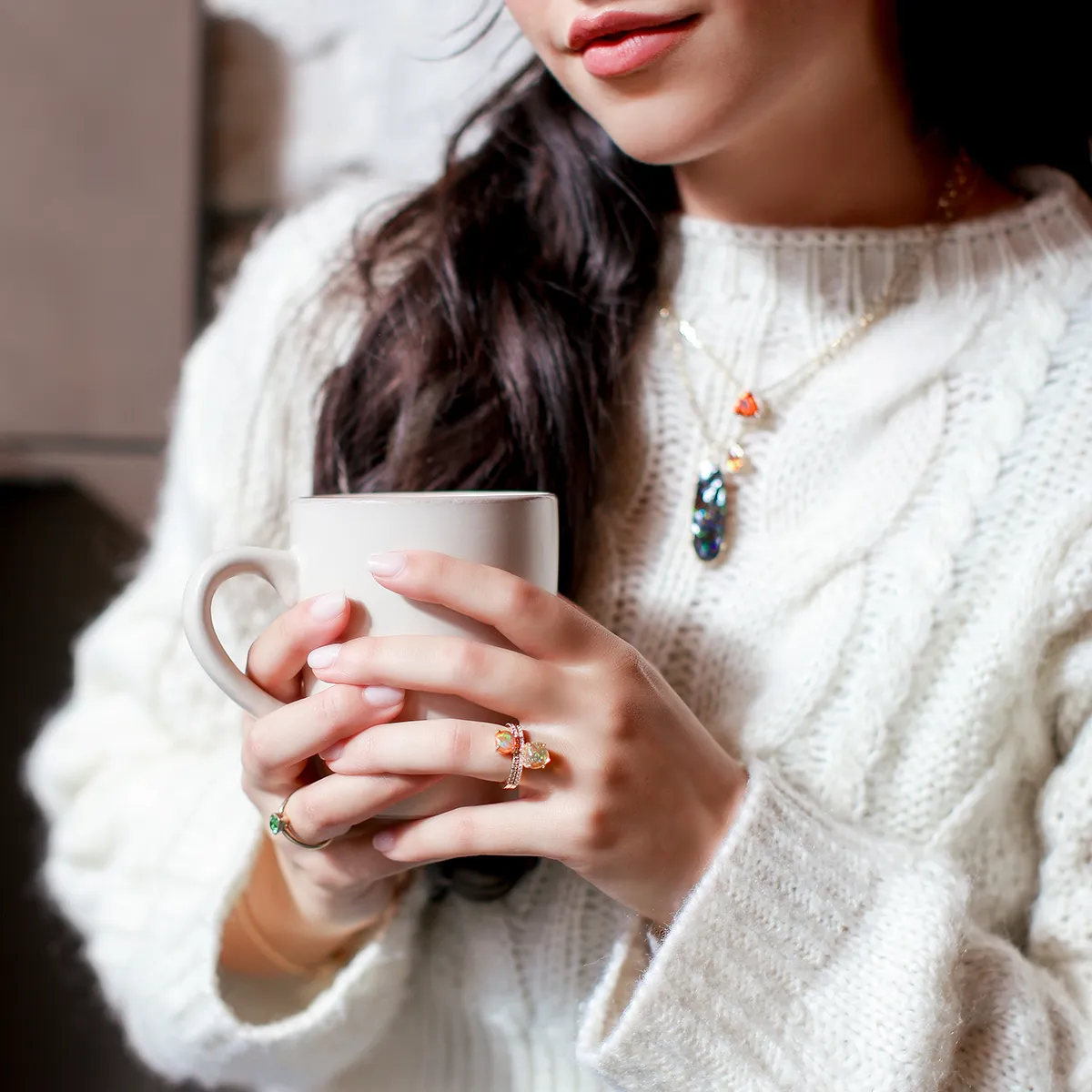 A woman in a white sweater holds a mug, wearing gemstone jewelry.