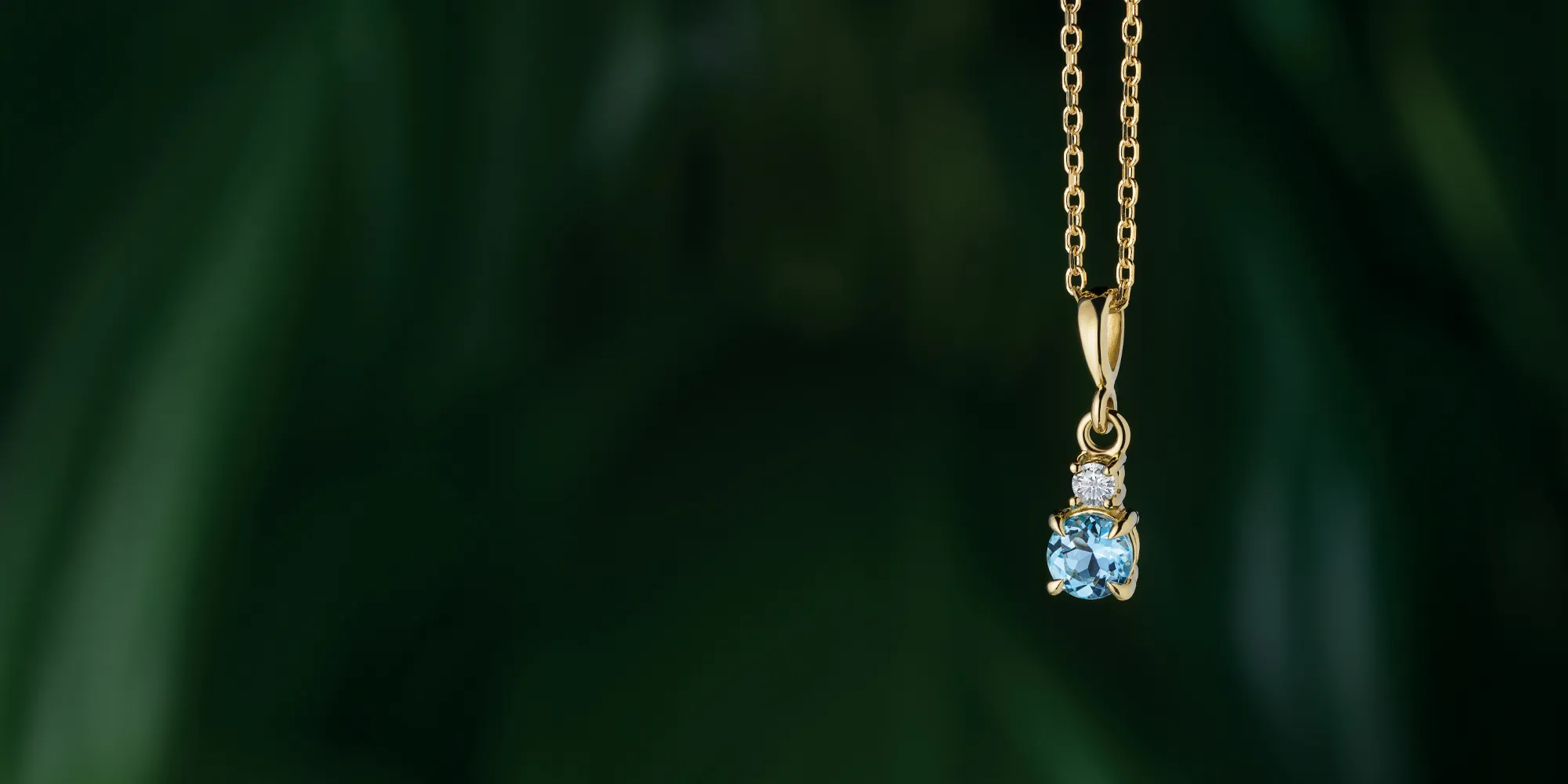 Gold chain with a blue gemstone pendant on a dark green background. Click to learn about custom jewelry at J. Anthony Jewelers.