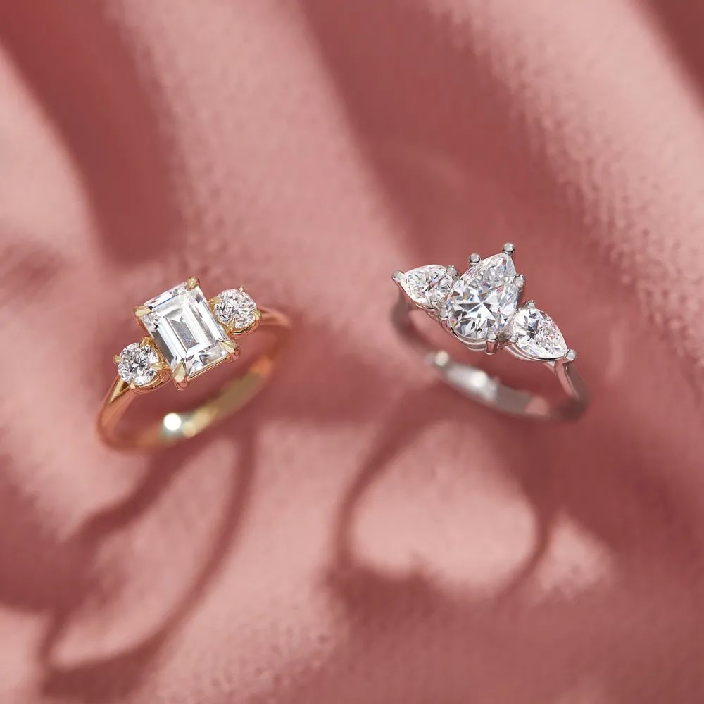 Two elegant engagement rings on a pink background, showcasing a diamond and emerald cut. Visit J. Anthony Jewelers in Neenah.
