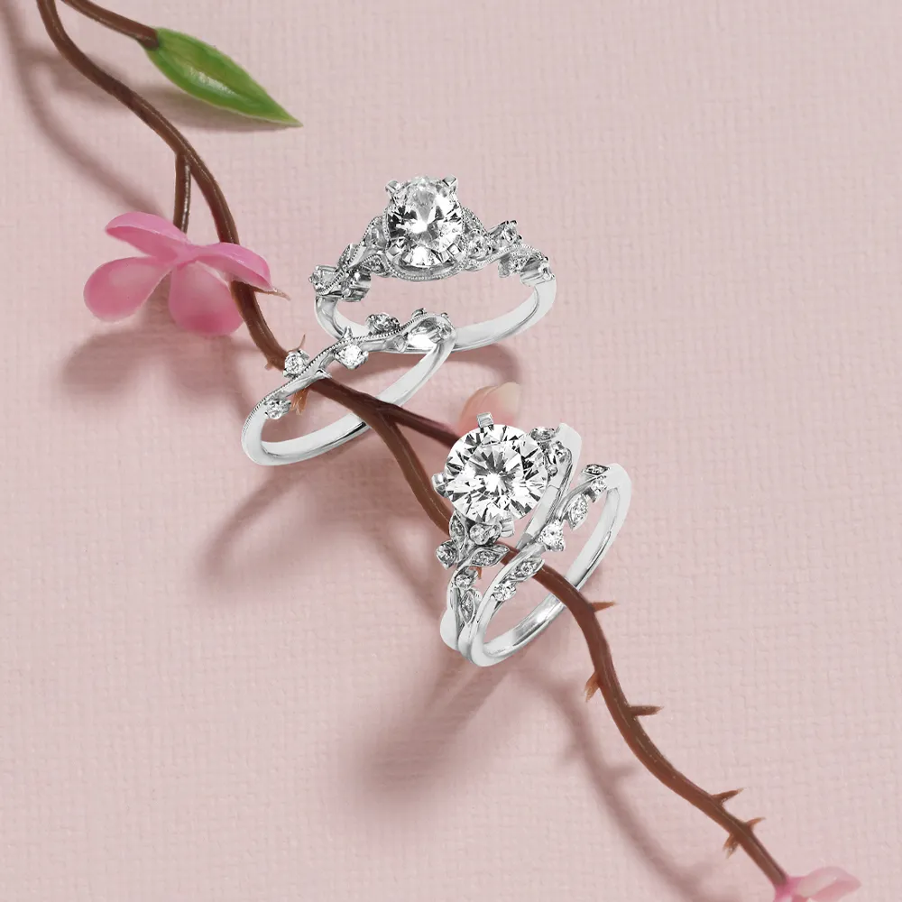 Two diamond engagement rings on a branch with pink flowers on a textured pink background.