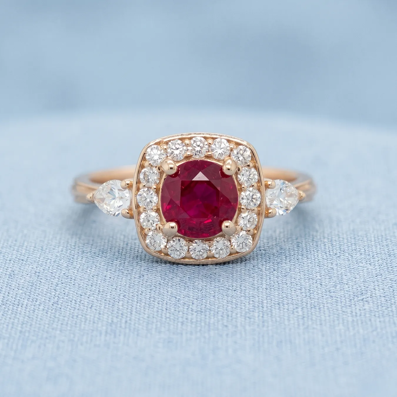A gold ring with a central red gemstone surrounded by small diamonds on a blue fabric background.
