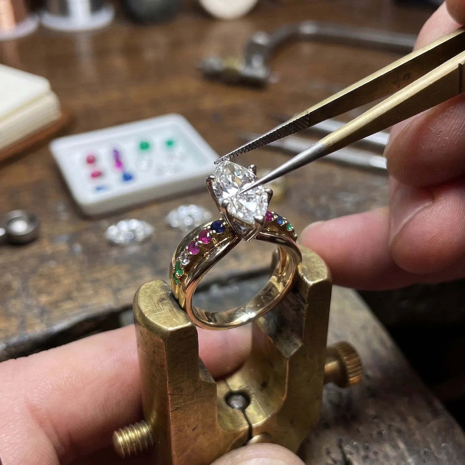 Jeweler setting a large diamond on a gold ring with colorful gemstones using tweezers.