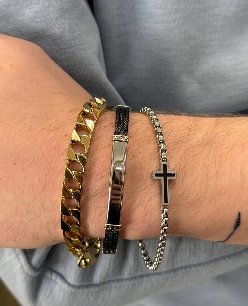 Three bracelets on a wrist: gold chain, black and silver band, and silver chain with a cross. Click to shop men's jewelry.