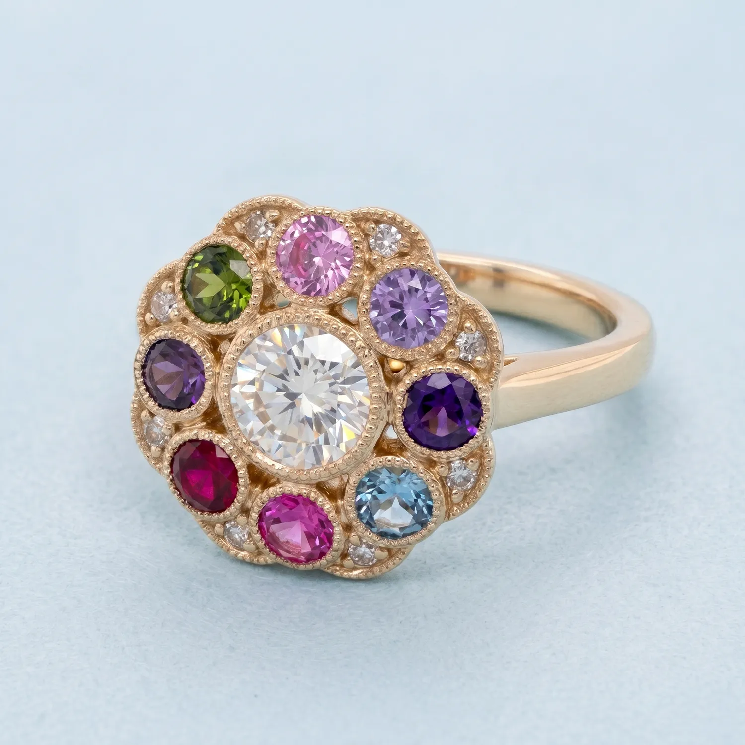 A gold ring with a large central diamond, surrounded by colorful gemstones in a floral pattern, on a light blue background.